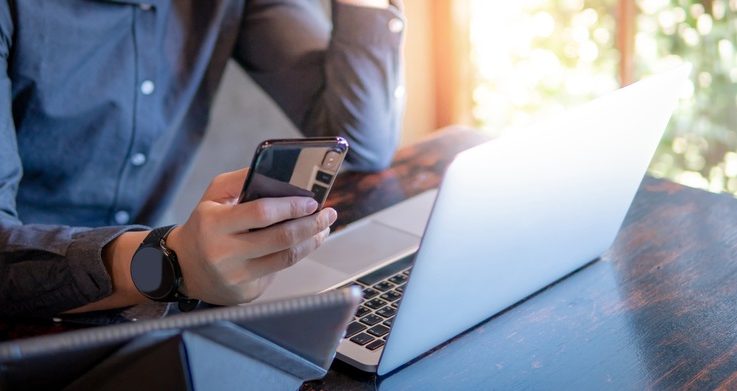 male hand holding smart phone in front of computer